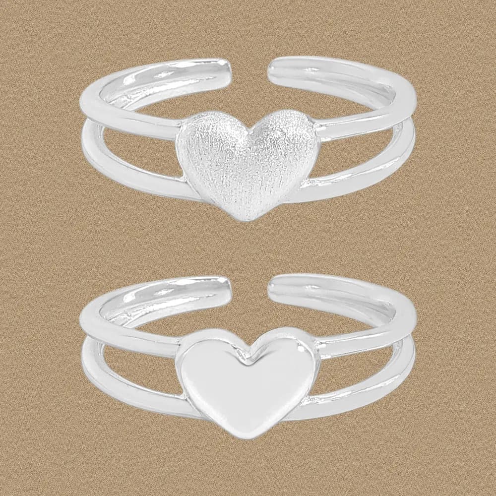 Two slim sterling silver heart rings stacked together, one textured and one smooth, on a warm beige background.