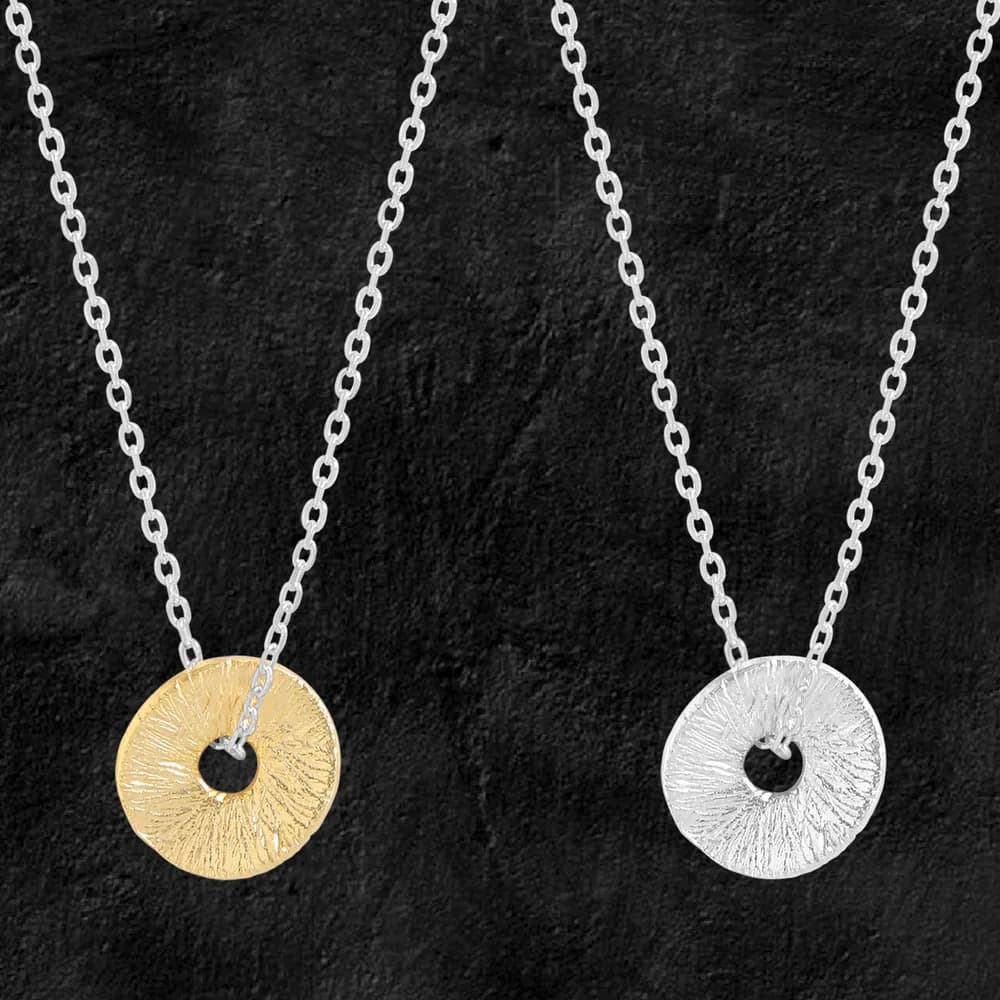 Two-tone textured circular disc pendants with central holes hanging on silver chains against a dark background.