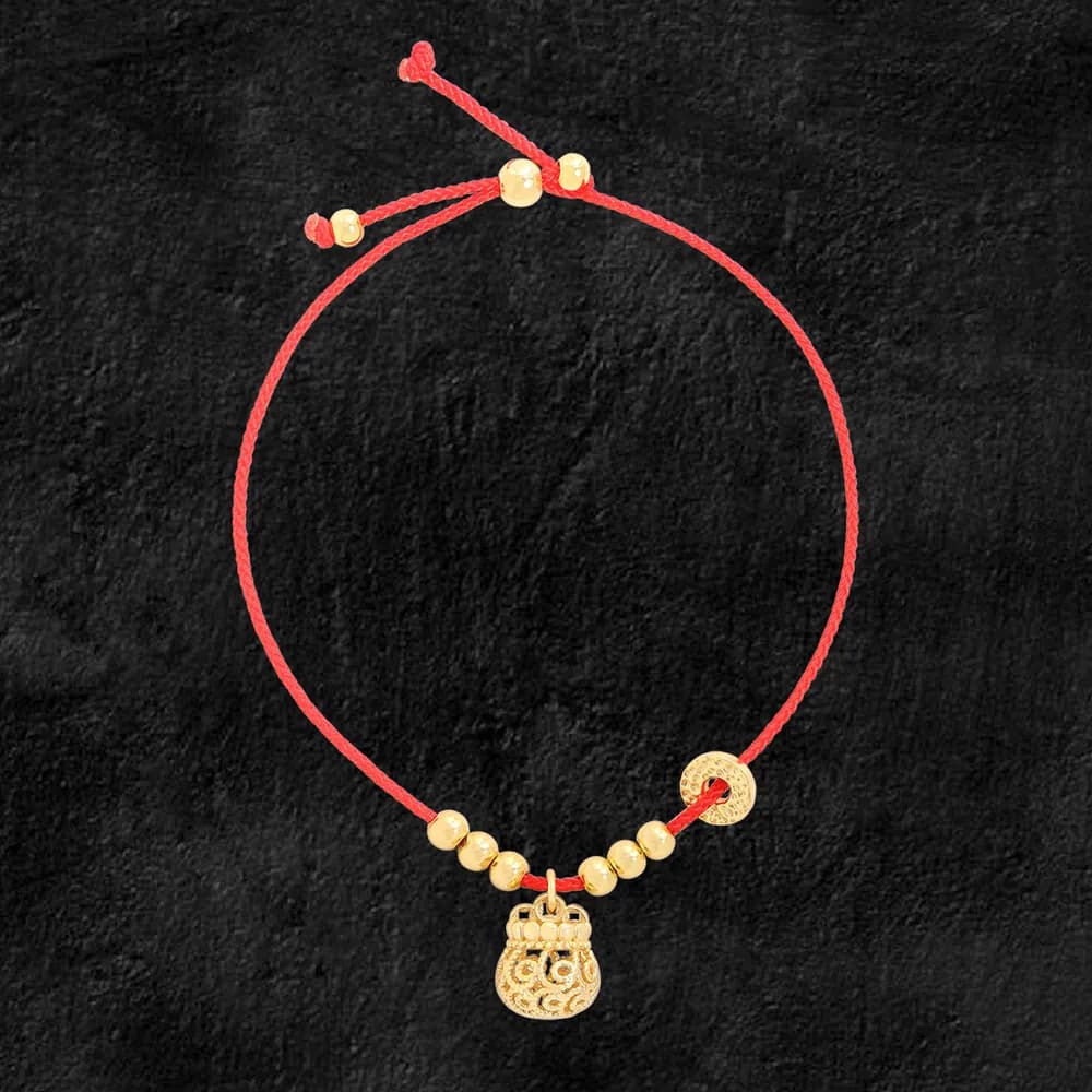 Red cord bracelet with gold-toned beads and a filigree pouch charm against a dark textured background.