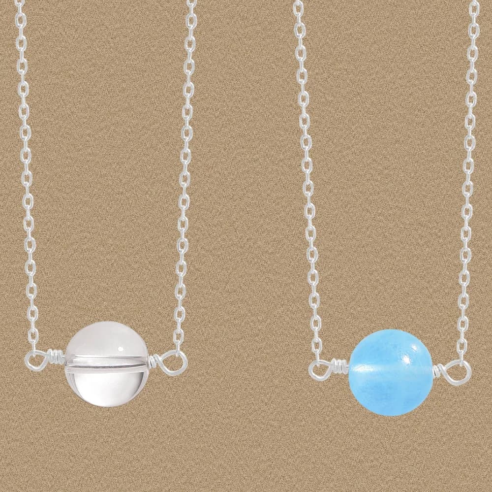 Two delicate silver chain necklaces each featuring a smooth enamel sphere pendant in white and blue on a sandy background.